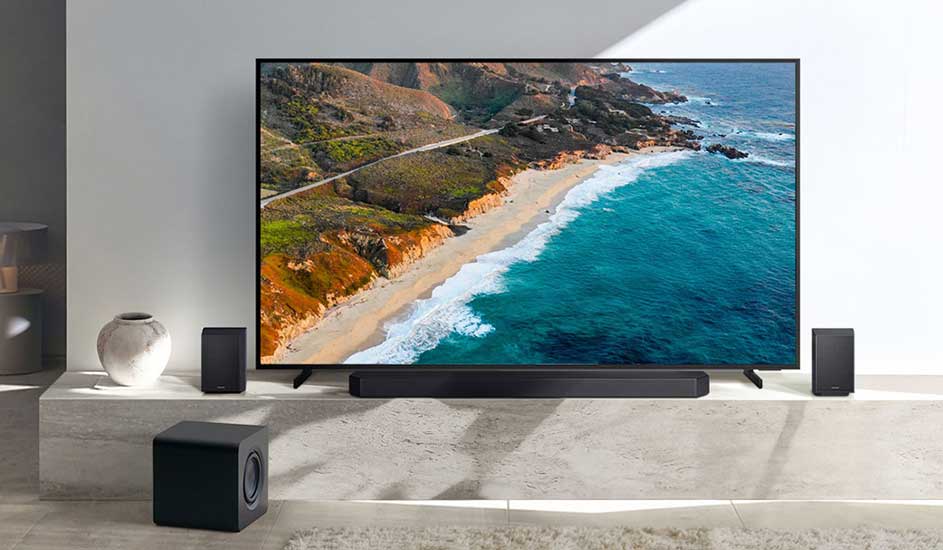Une télévision Samsung est au centre avec une image de nature à l’écran, entourée d’une barre de son Samsung, d’un caisson de basses et de deux enceintes dans un salon moderne.