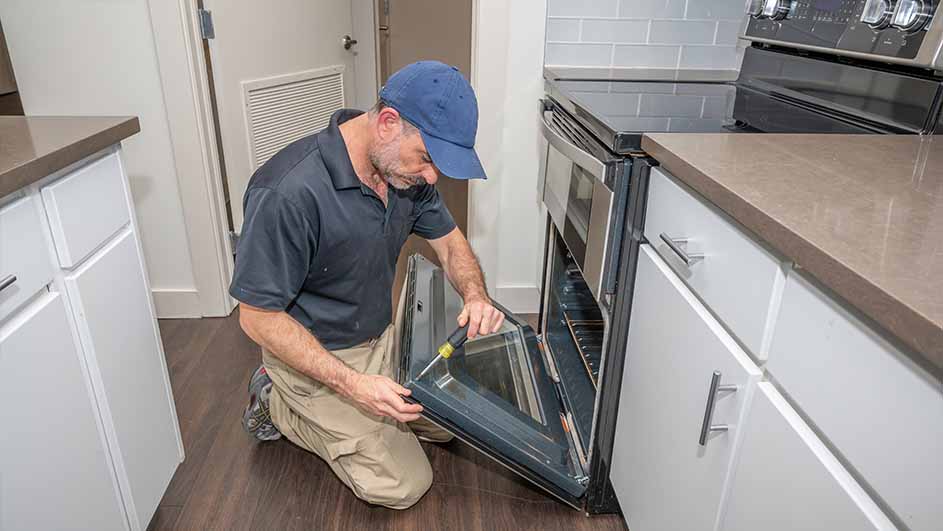 Een man knielt voor een open oven in een keuken met witte kasten en een aanrechtblad. Hij gebruikt een schroevendraaier.
