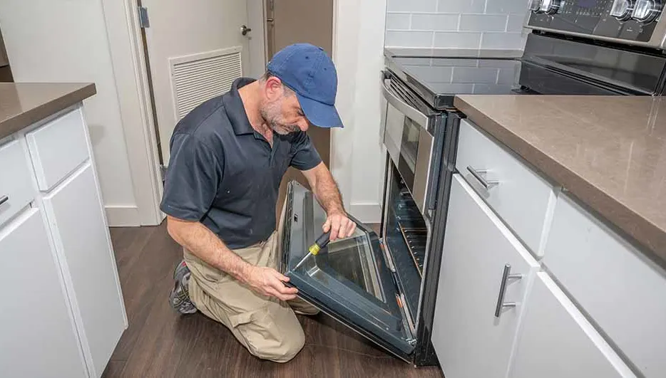 Een man knielt voor een open oven in een keuken met witte kasten en een aanrechtblad. Hij gebruikt een schroevendraaier.