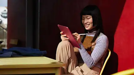 Une tablette rose est utilisée avec un stylet sur une chaise jaune devant un mur rouge, près d'une table jaune.