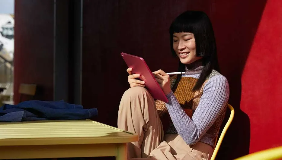Une tablette rose est utilisée avec un stylet sur une chaise jaune devant un mur rouge, près d'une table jaune.