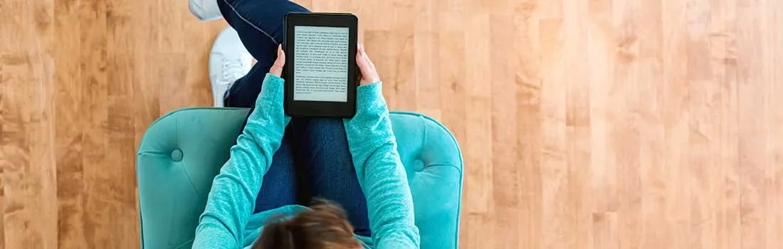 An e-reader is held above a teal chair. The chair sits on a light-colored wooden floor.