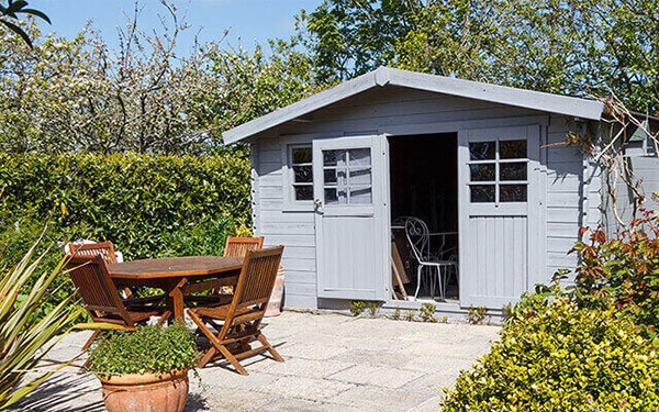 Une remise grise avec une porte ouverte est située sur une terrasse en pierre, avec une table et des chaises en bois.