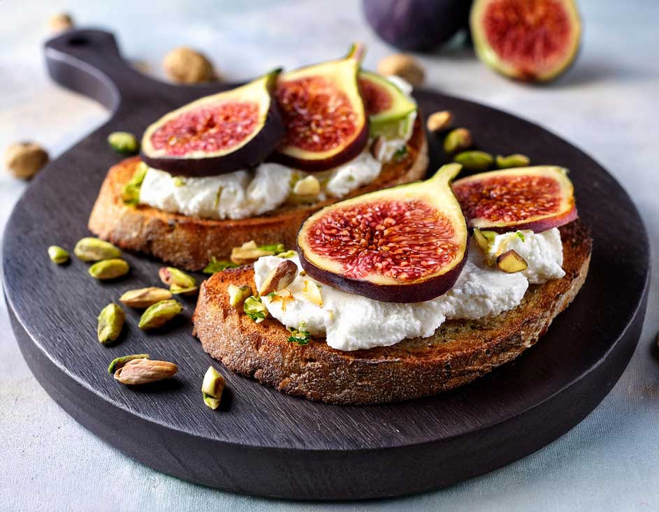 Deux tartines sur planche brune : pain grillé, fromage blanc, figues coupées et pistaches hachées. Deux figues entières en arrière-plan.