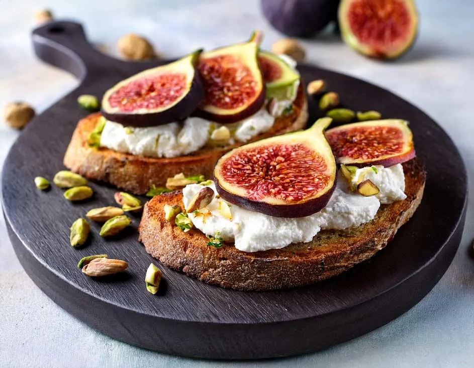 Deux tartines sur planche brune : pain grillé, fromage blanc, figues coupées et pistaches hachées. Deux figues entières en arrière-plan.