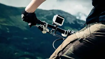 Close-up of a bike handlebar with a camera attached, mountains in background. Gloved hand grips the bar, earthy-toned pants.