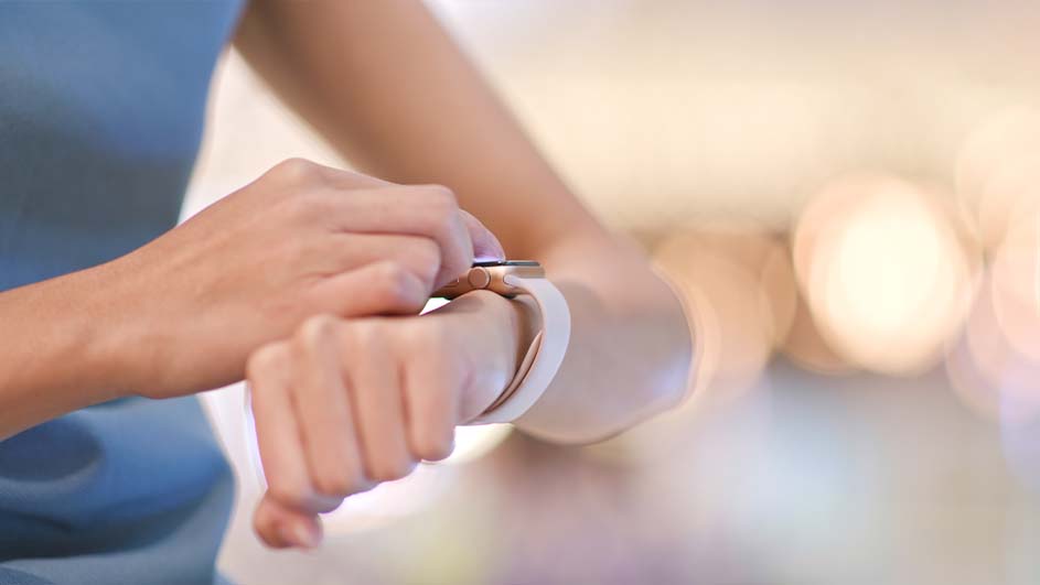 Een pols met een witte smartwatch wordt aangeraakt, met een blauw shirt en een zachte, lichte achtergrond.