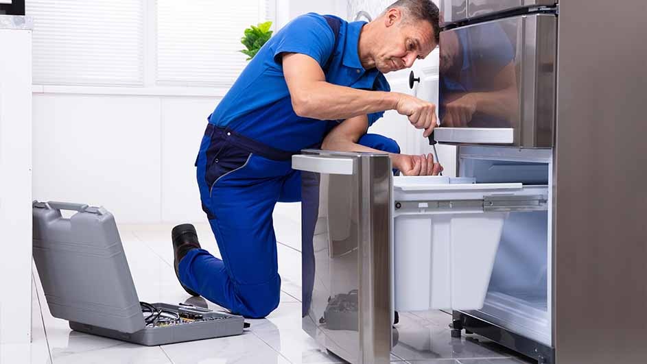A person in blue workwear is repairing a stainless steel refrigerator with a screwdriver. A grey toolbox sits nearby.