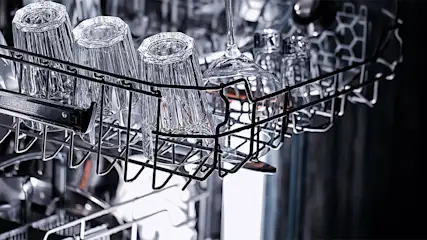 Dans un lave-vaisselle, un panier métallique noir contient des verres transparents, avec des couverts en arrière-plan.
