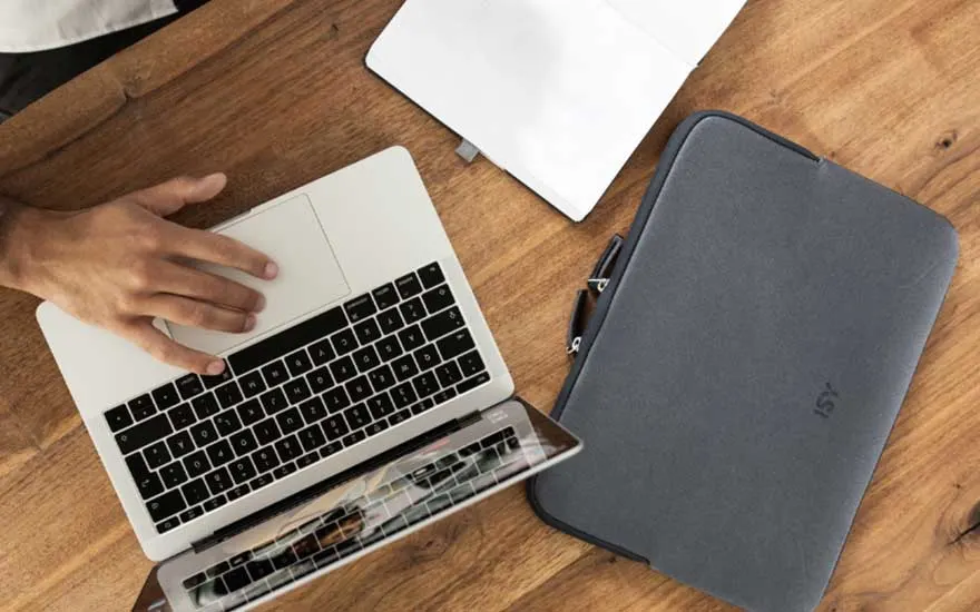 Ordinateur portable avec main sur clavier à côté d'une housse grise sur table en bois. Configuration moderne pour télétravail.