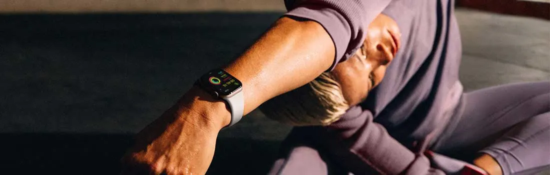 Une personne porte un survêtement violet et une montre intelligente avec un bracelet gris, assise sur une surface grise.
