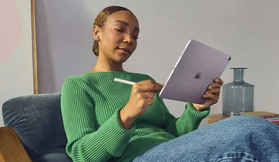 Een persoon zit op een bank en gebruikt een stylus op een tablet, met een groene trui, spijkerbroek en een vaas op tafel ernaast.