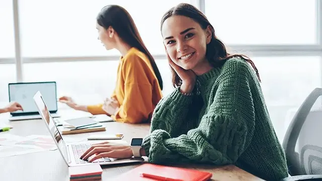 Een lachende vrouw in een groene trui zit aan een bureau met laptop, terwijl een collega achter haar werkt in een lichte kantoorruimte.