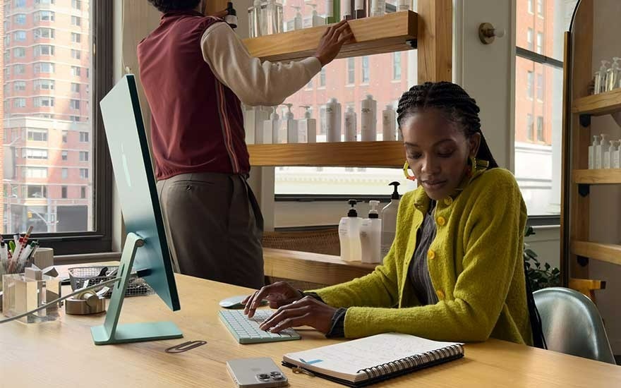An office scene with a light wood desk holding a teal computer, keyboard, and notebook. Shelves with bottles line the wall. Outside, a city building is visible.
