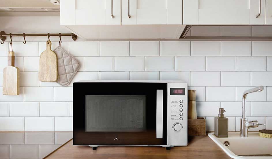 Un micro-ondes noir et blanc sur un comptoir de cuisine en bois, avec carrelage blanc en arrière-plan. Étagères et ustensiles visibles.
