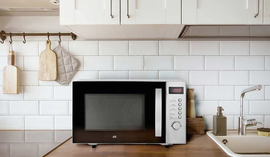 Un micro-ondes noir et blanc sur un comptoir de cuisine en bois, avec carrelage blanc en arrière-plan. Étagères et ustensiles visibles.