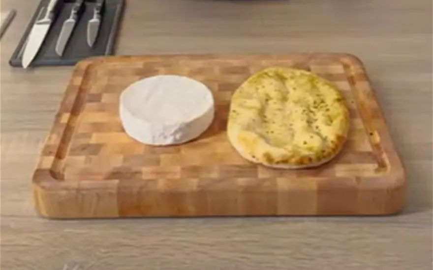 Op een houten plank ligt een witte kaas naast een plat brood. Op de achtergrond staan messen.
