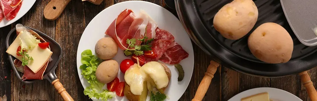 Een bovenaanzicht toont ingrediënten voor raclette, zoals aardappelen, vleeswaren en kaas, op een rustieke houten tafel.