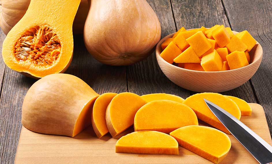 Des courges butternut, entières, coupées et en dés, sur une planche à découper en bois, avec un couteau, sur une table en bois.