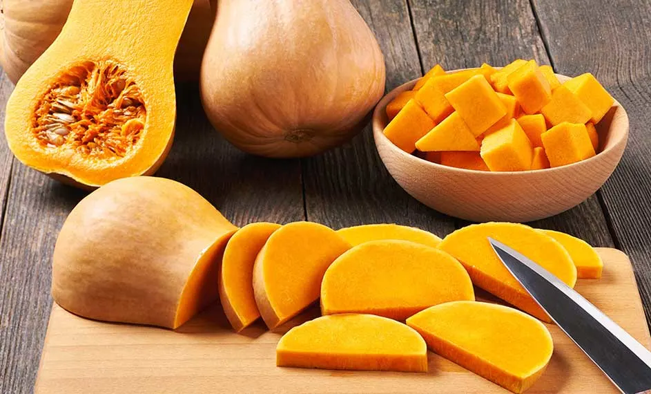 Op een houten tafel liggen butternutpompoenen, zowel hele als gesneden, naast een houten kom met pompoenblokjes en een mes.