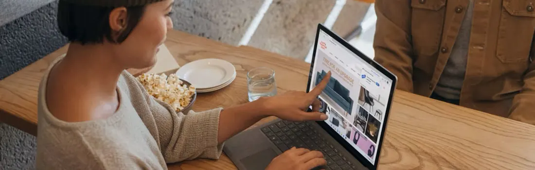 Op een houten tafel staat een grijze laptop met een website. Er staat ook een kom popcorn, een stapel borden en een glas water.