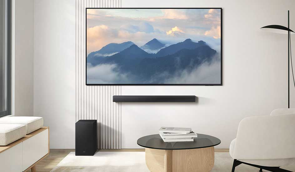 Un téléviseur LG affiche des montagnes dans les nuages au mur. En dessous, une barre de son LG et un caisson de basses dans un salon moderne et lumineux.