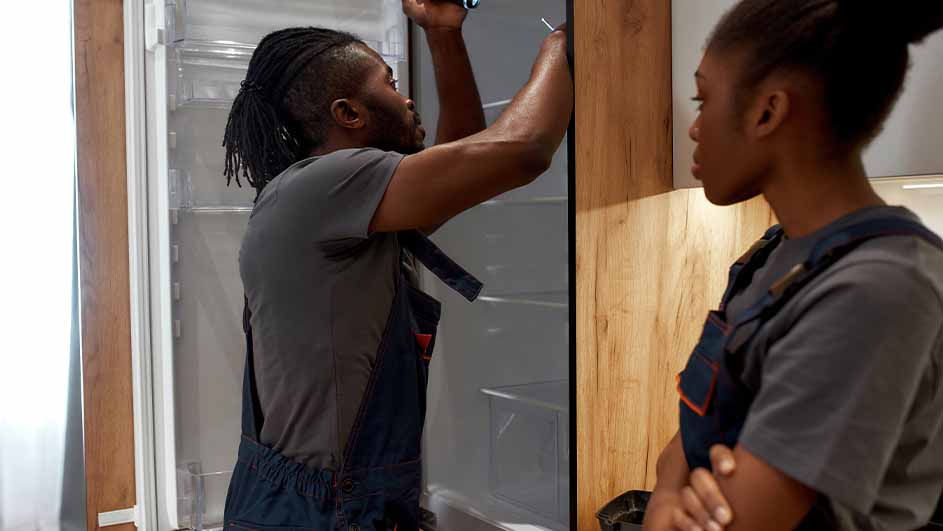 Twee mensen in grijze shirts en blauwe overalls. Een koelkast met open deur en houten panelen zijn zichtbaar.