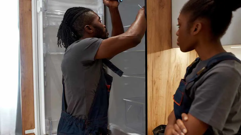 Twee mensen in grijze shirts en blauwe overalls. Een koelkast met open deur en houten panelen zijn zichtbaar.