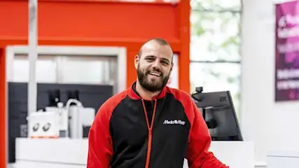 Een zwarte en rode jas met MediaMarkt logo voor een kassa met computerscherm en oranje accenten.