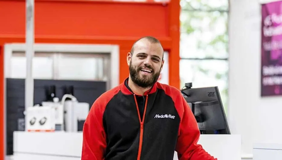 Een zwarte en rode jas met MediaMarkt logo voor een kassa met computerscherm en oranje accenten.