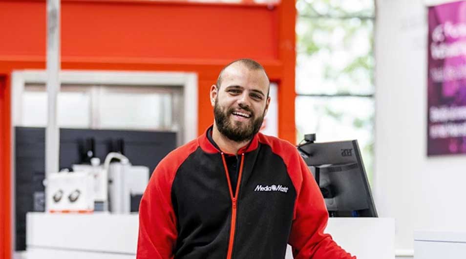 Un comptoir blanc avec un ordinateur et une caisse enregistreuse devant un mur rouge et blanc. Veste rouge et noire avec le logo MediaMarkt.