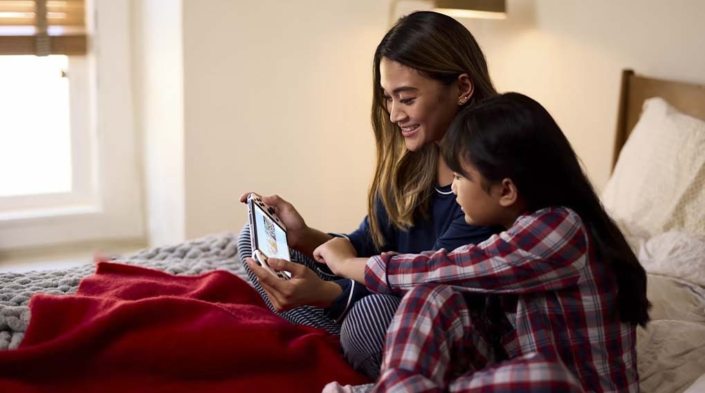 Sur un lit avec une couverture rouge, une personne et un enfant regardent un appareil électronique portable.