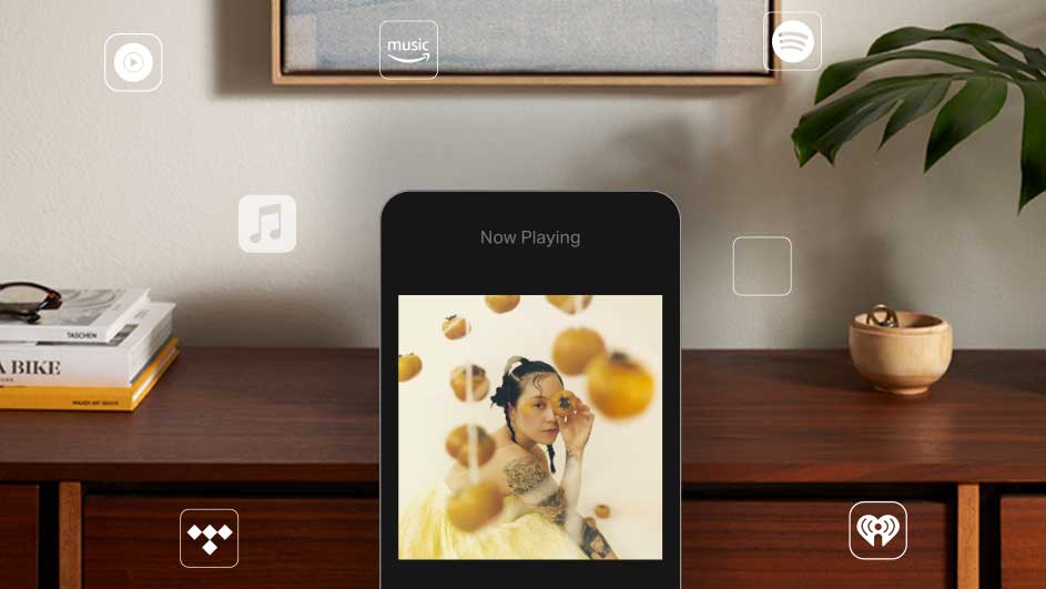 Een smartphone toont een afspeelscherm met albumhoes, op een houten kast met boeken en decoratieve objecten.