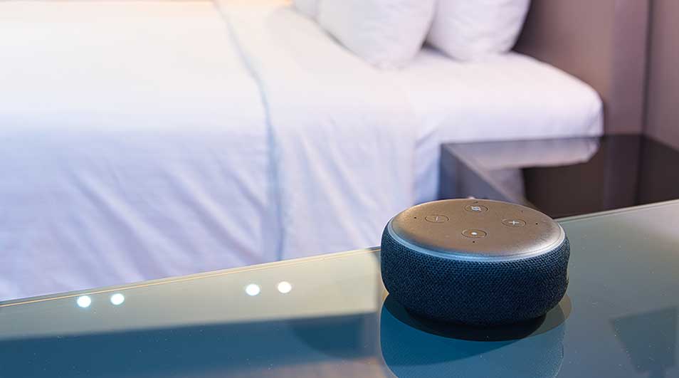 Une enceinte intelligente ronde noire sur une table en verre, avec un lit blanc et une table de chevet en arrière-plan.