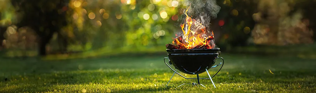 Un barbecue noir allumé avec des flammes et de la fumée sur une pelouse verte. Arrière-plan flou avec des arbres.