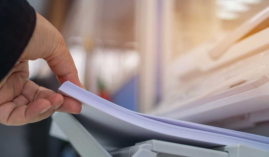 Een hand pakt een stapel wit papier uit een printer. De achtergrond is vaag.