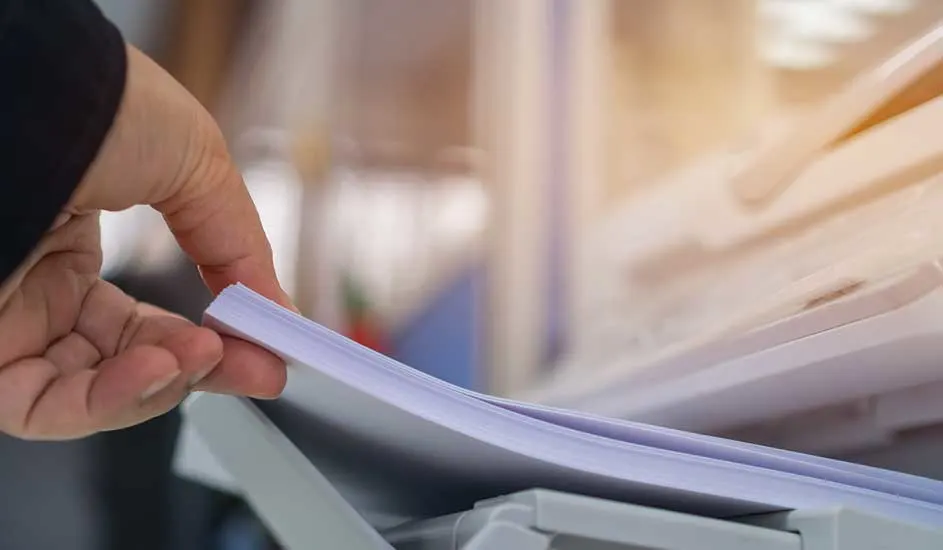 Een hand pakt een stapel wit papier uit een printer. De achtergrond is vaag.