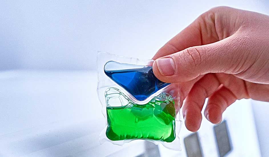 Une main tient une capsule de lessive transparente contenant un liquide bleu et vert, sur un fond blanc.
