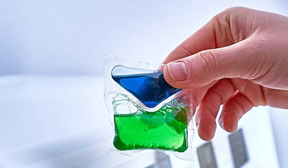 Une main tient une capsule de lessive transparente contenant un liquide bleu et vert, sur un fond blanc.