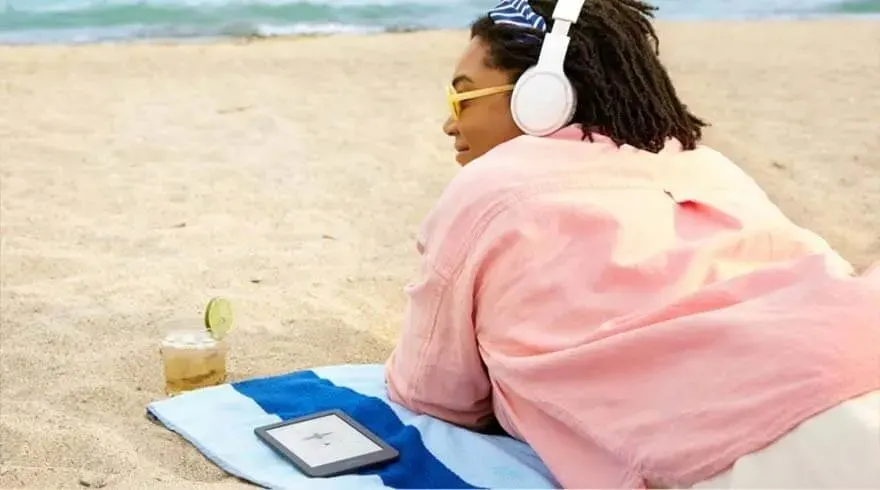 Een strandscène met een drankje, een e-reader en een blauw-wit gestreepte handdoek op zand. De zee is zichtbaar op de achtergrond.