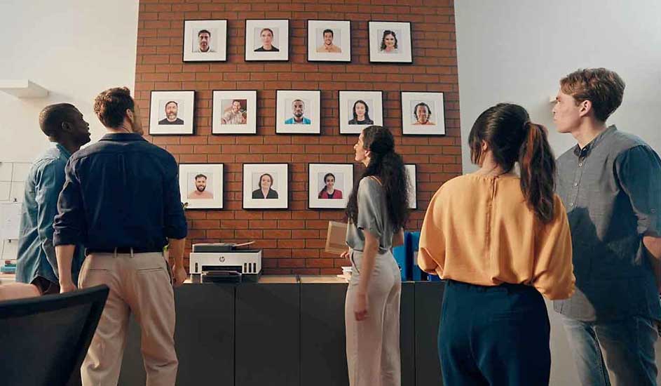 Des personnes regardent des photos encadrées sur un mur en briques, au-dessus d'un bureau avec une imprimante.