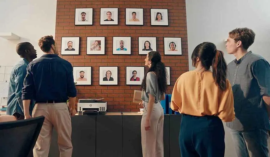 Des personnes regardent des photos encadrées sur un mur en briques, au-dessus d'un bureau avec une imprimante.