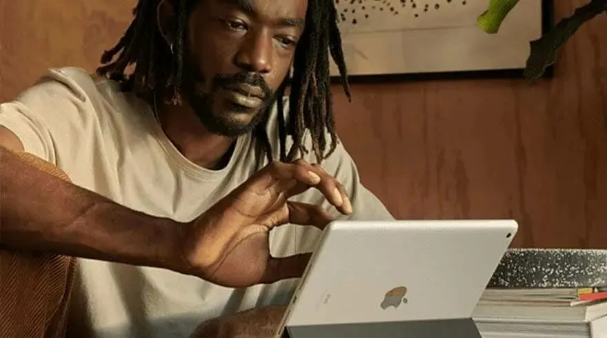 Un iPad argenté est utilisé sur une table. Un mur en bois et une plante sont visibles en arrière-plan.