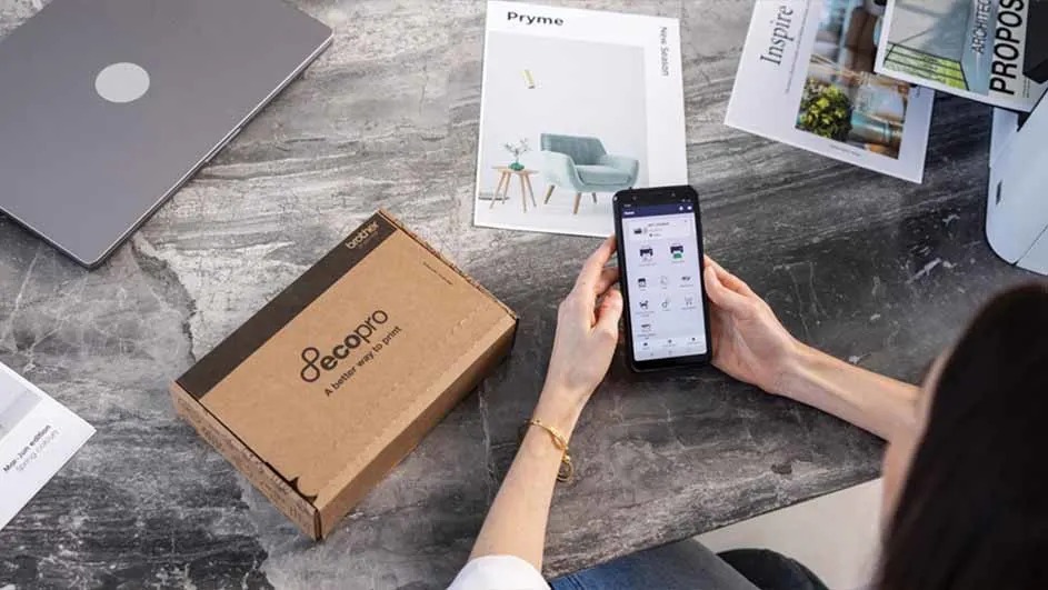 Een grijze laptop, een bruine doos, een telefoon in handen en brochures liggen op een grijze marmeren tafel.