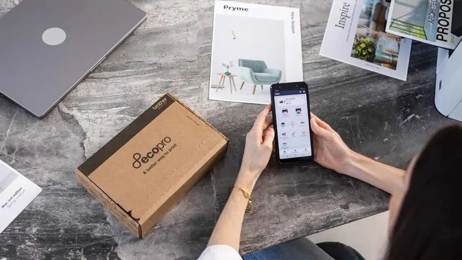 Een grijze laptop, een bruine doos, een telefoon in handen en brochures liggen op een grijze marmeren tafel.