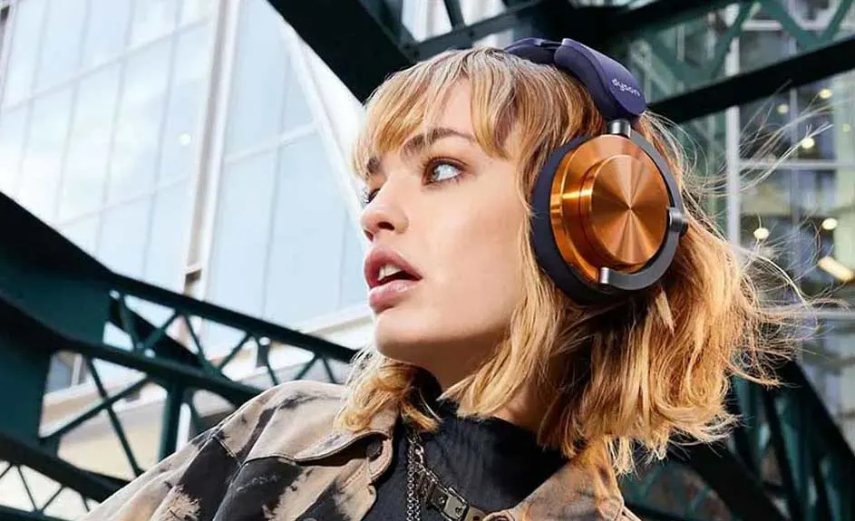 Blond haar met een donkerblauwe hoofdtelefoon met koperen oorschelpen. Achtergrond: gebouw en groene metalen structuur.