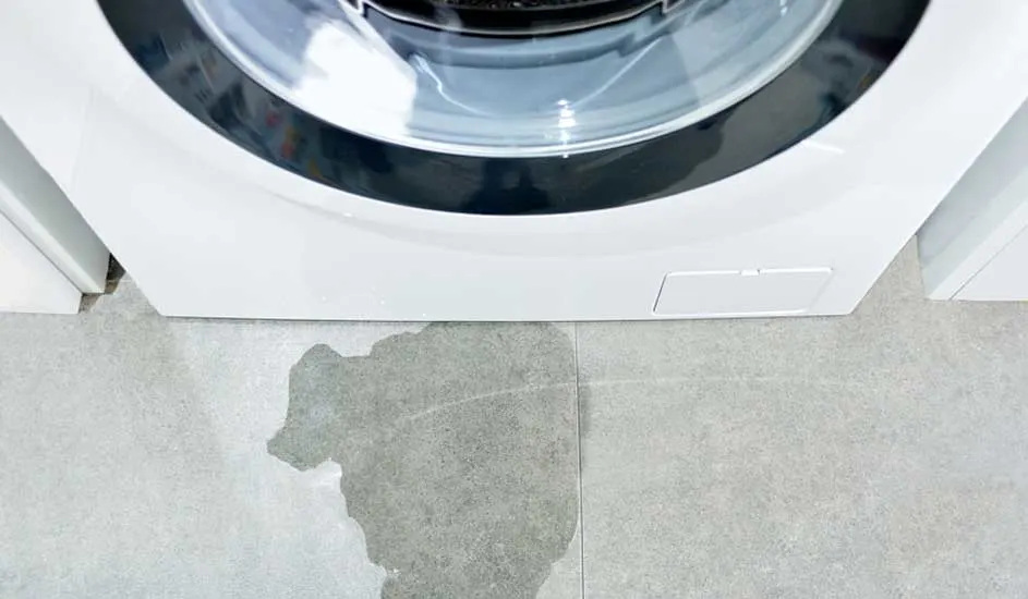 Vue rapprochée d'une machine à laver blanche avec une fuite d'eau sur un carrelage gris.