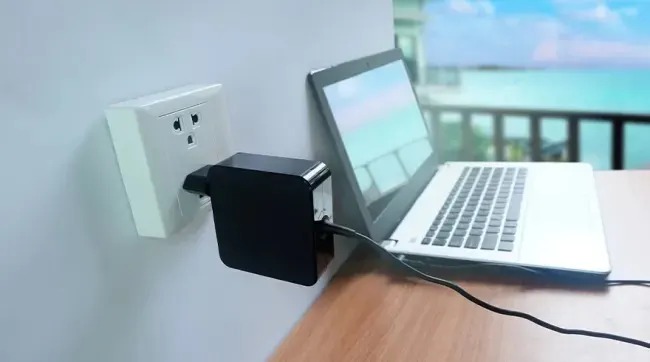 Un ordinateur portable blanc est branché sur une prise murale avec un adaptateur noir, avec vue sur la mer en arrière-plan.