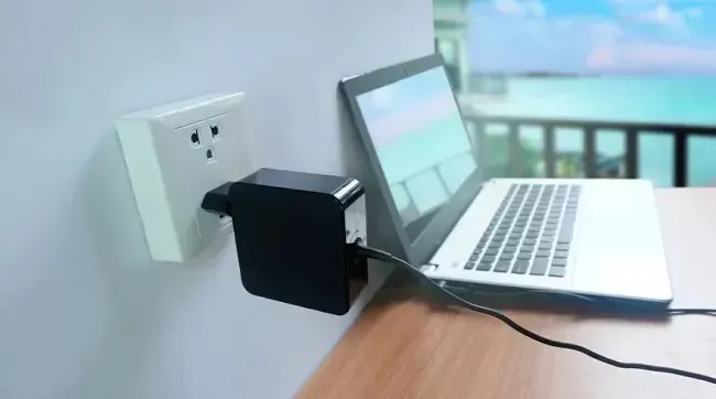 Un ordinateur portable blanc est branché sur une prise murale avec un adaptateur noir, avec vue sur la mer en arrière-plan.