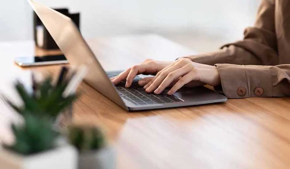 Une personne utilise un ordinateur portable sur une table en bois, avec un téléphone, un pot à crayons et une plante en arrière-plan.